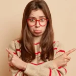 تصویر زن جوانی با عینک قرمز و پلیور بافتنی راه راه که با حالتی سردرگم و مردد، با انگشتانش به دو جهت مخالف (چپ و راست) اشاره می‌کند.