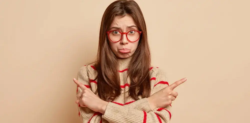 تصویر زن جوانی با عینک قرمز و پلیور بافتنی راه راه که با حالتی سردرگم و مردد، با انگشتانش به دو جهت مخالف (چپ و راست) اشاره می‌کند.